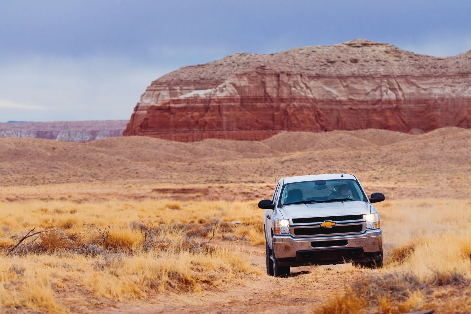 White pickup on a dirt track in dry grass below a red banded mesa