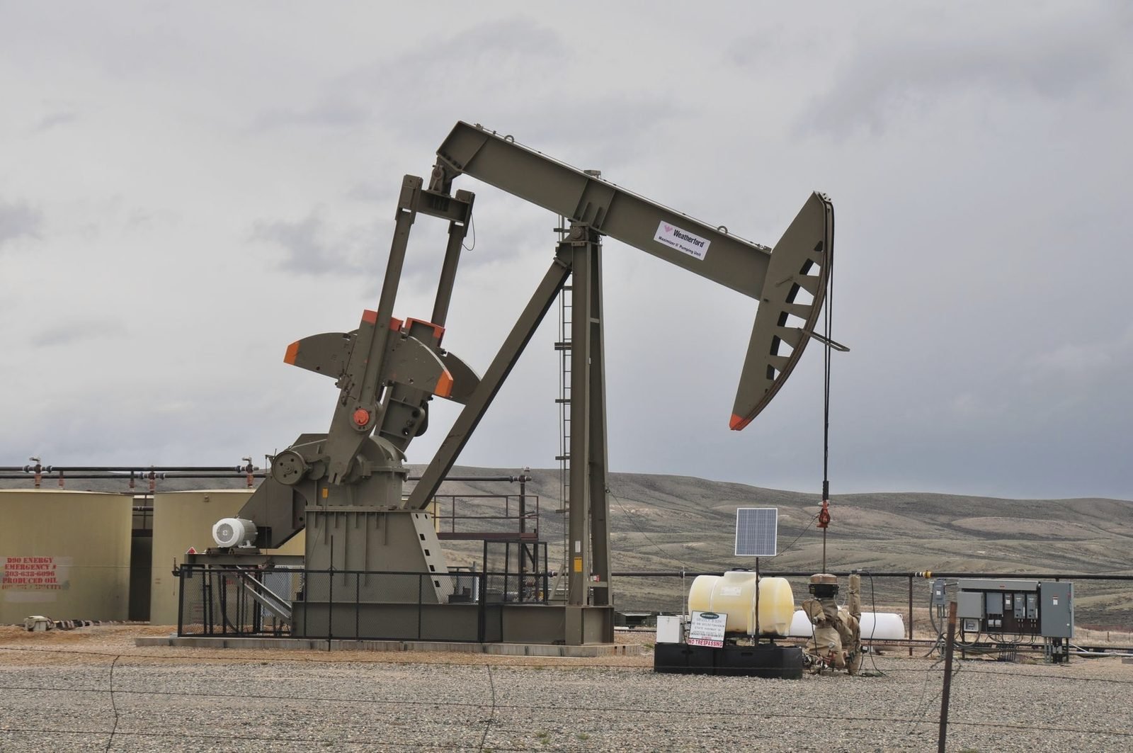 Pumpjack inside a fenced site with tanks and equipment under grey sky
